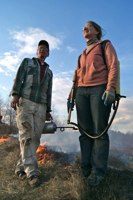 A man and a woman each holding field burning equipment with a portion of dried grass on fire behind them. 