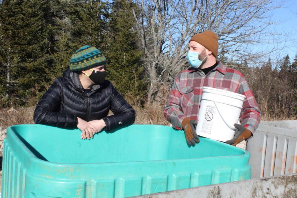 Two people leaning up against a large square plastic bin used for compost.