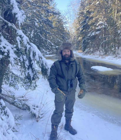 A man dressed in a Maine Inland Fisheries and Wildlife uniform and a fur hat standing next to a partially frozen stream in a forested area in winter.